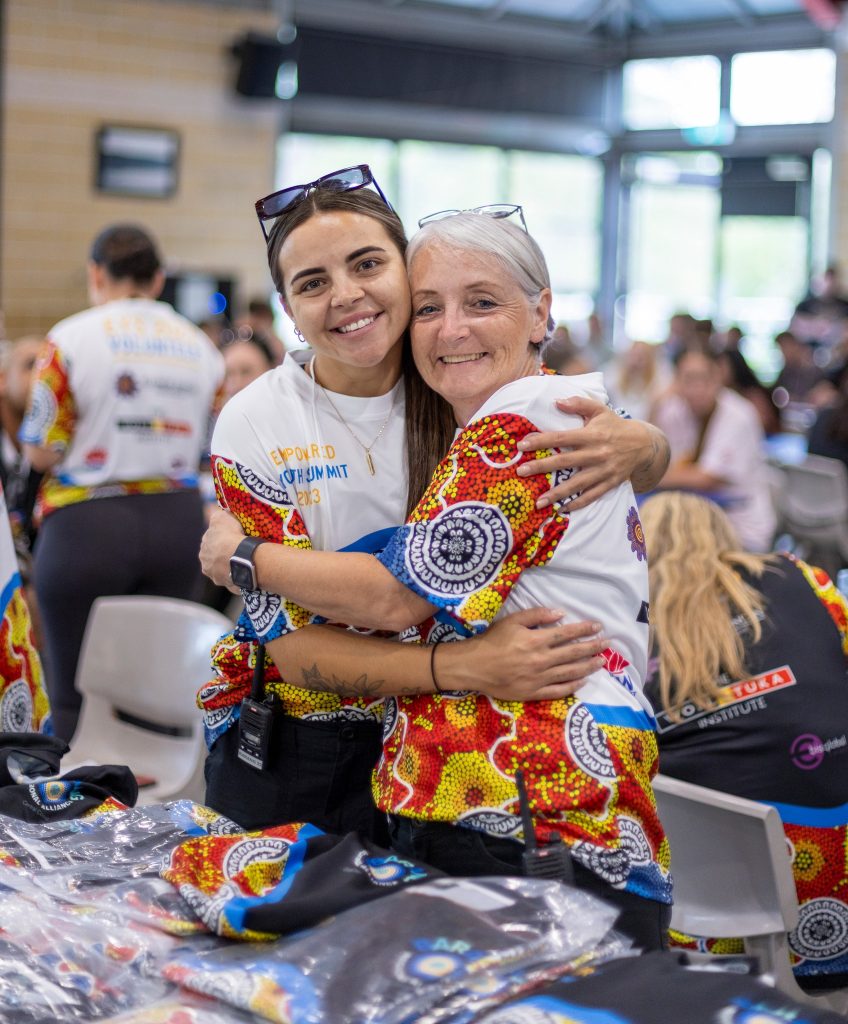 Two women embrace and smile at the camera, wearing matching, colorful shirts featuring intricate dot patterns. Behind them, others are seated and engaged in activities, suggesting a well-lit indoor community event. This snapshot captures the vibrant spirit of the Weekend of Culture.